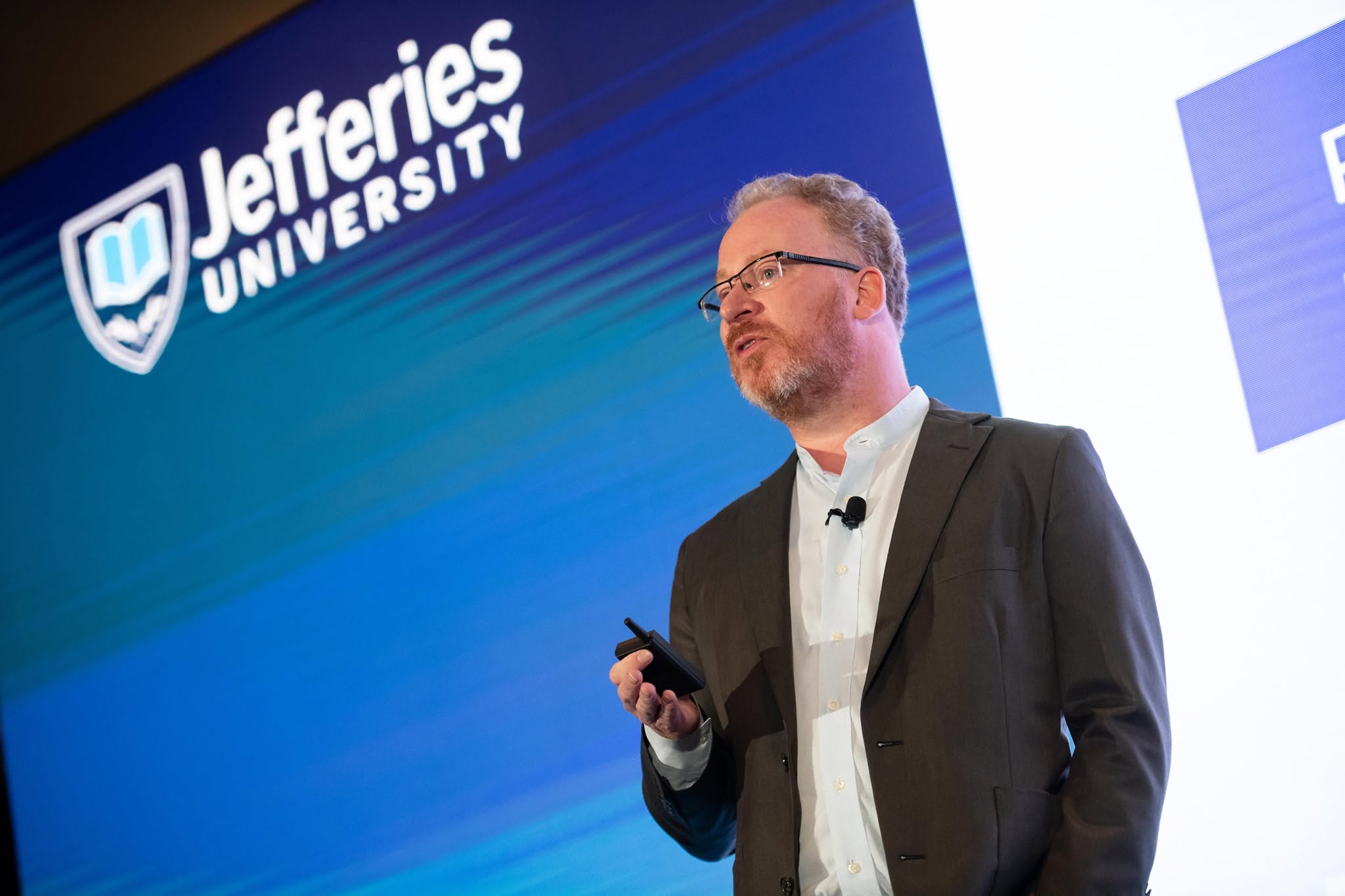 Robert Smithson speaking on stage in front of a large screen displaying the Jefferies University logo.